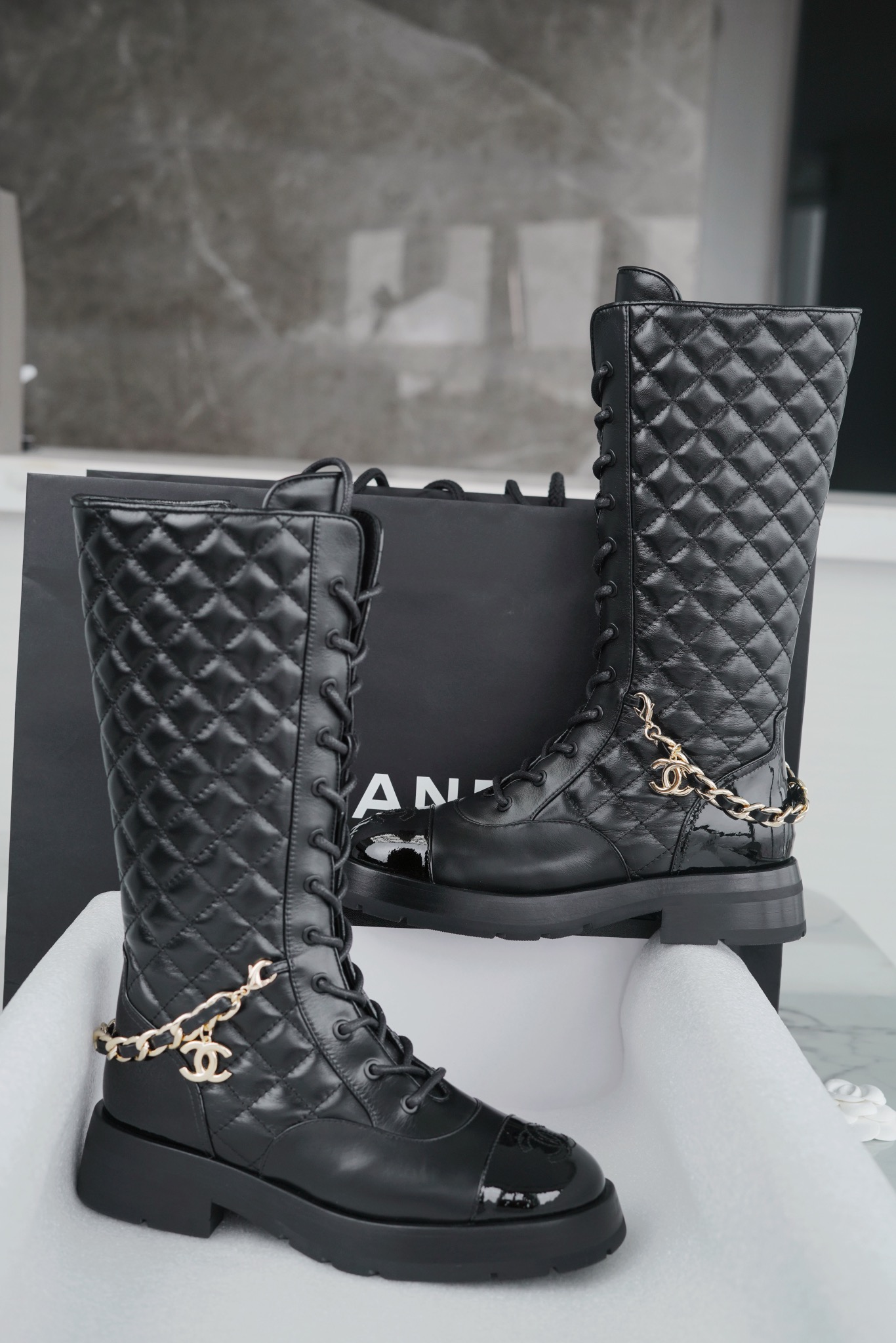 Chanel  23b  Chain Long Martin Boots: Crafted from black oil leather and black patent leather, with a leather combination sole. Classic diamond-pattern stitching and chain elements intertwine on the vamp