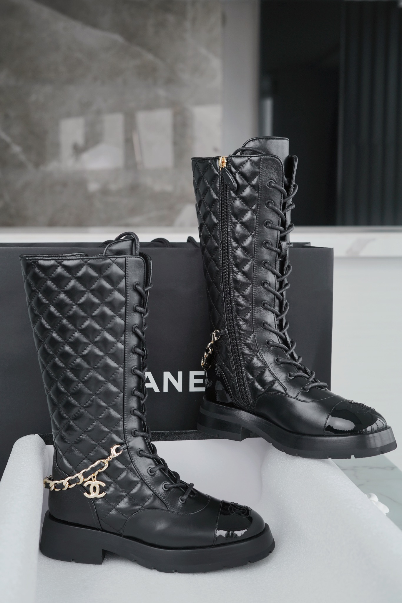 Chanel  23b  Chain Long Martin Boots: Crafted from black oil leather and black patent leather, with a leather combination sole. Classic diamond-pattern stitching and chain elements intertwine on the vamp