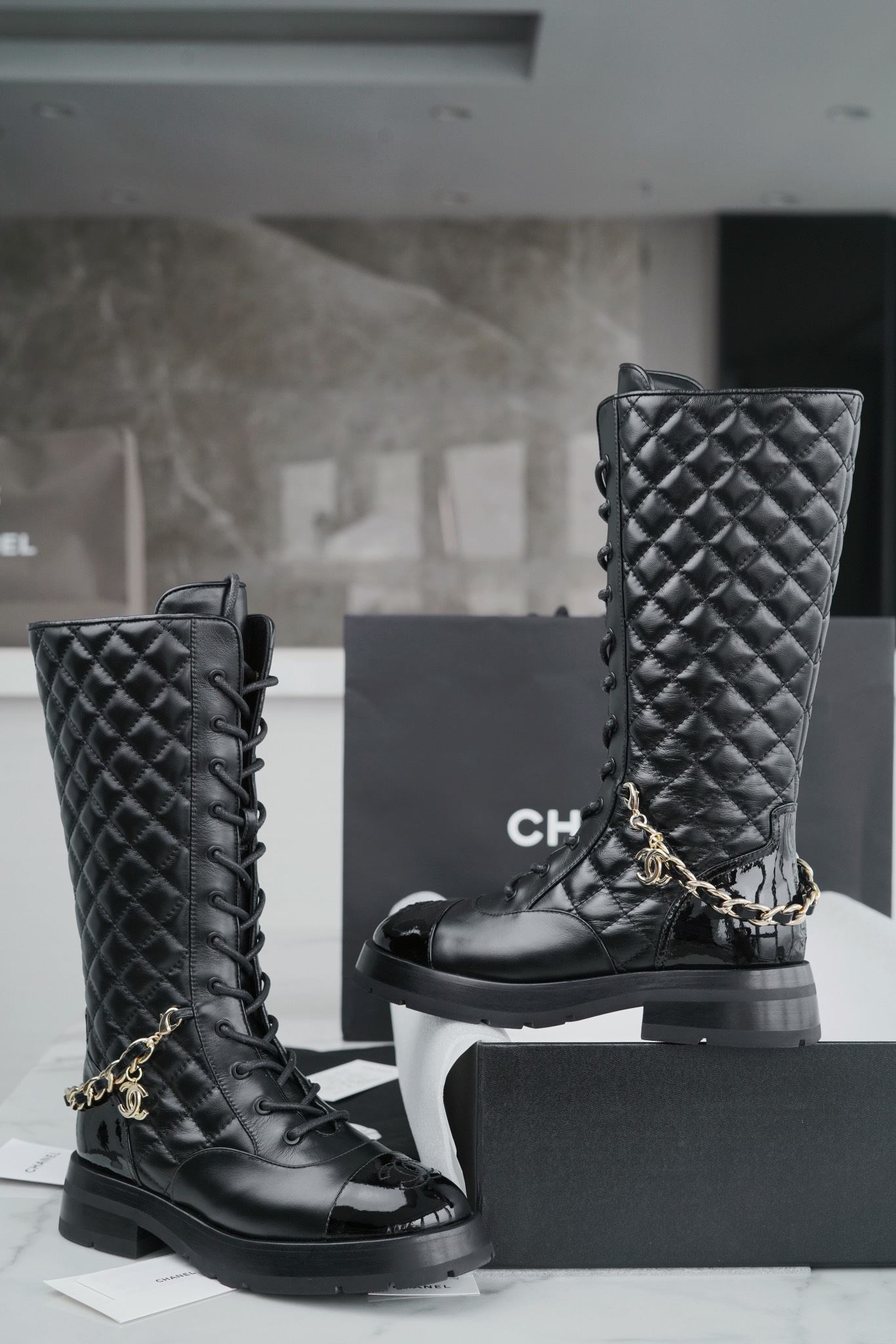 Chanel  23b  Chain Long Martin Boots: Crafted from black oil leather and black patent leather, with a leather combination sole. Classic diamond-pattern stitching and chain elements intertwine on the vamp