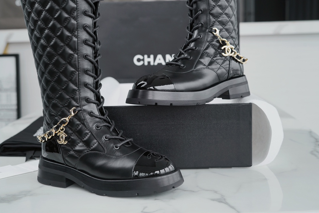 Chanel  23b  Chain Long Martin Boots: Crafted from black oil leather and black patent leather, with a leather combination sole. Classic diamond-pattern stitching and chain elements intertwine on the vamp