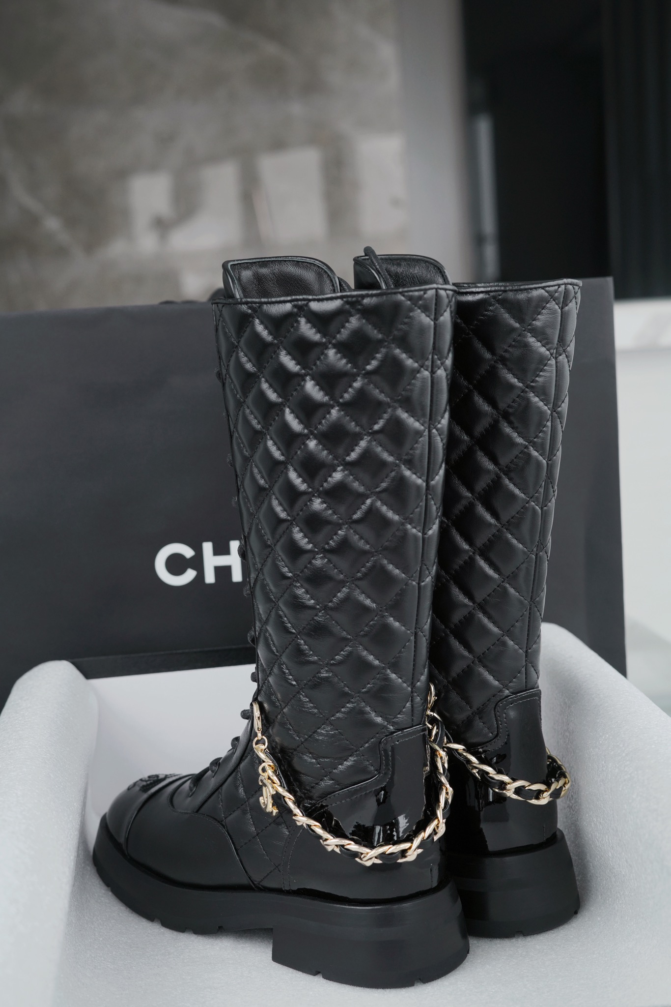 Chanel  23b  Chain Long Martin Boots: Crafted from black oil leather and black patent leather, with a leather combination sole. Classic diamond-pattern stitching and chain elements intertwine on the vamp