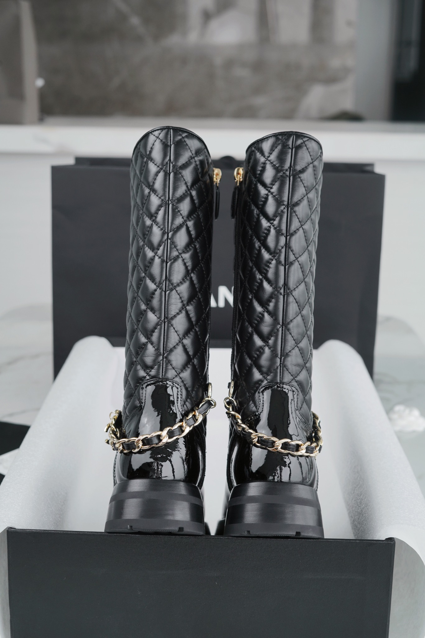 Chanel  23b  Chain Long Martin Boots: Crafted from black oil leather and black patent leather, with a leather combination sole. Classic diamond-pattern stitching and chain elements intertwine on the vamp