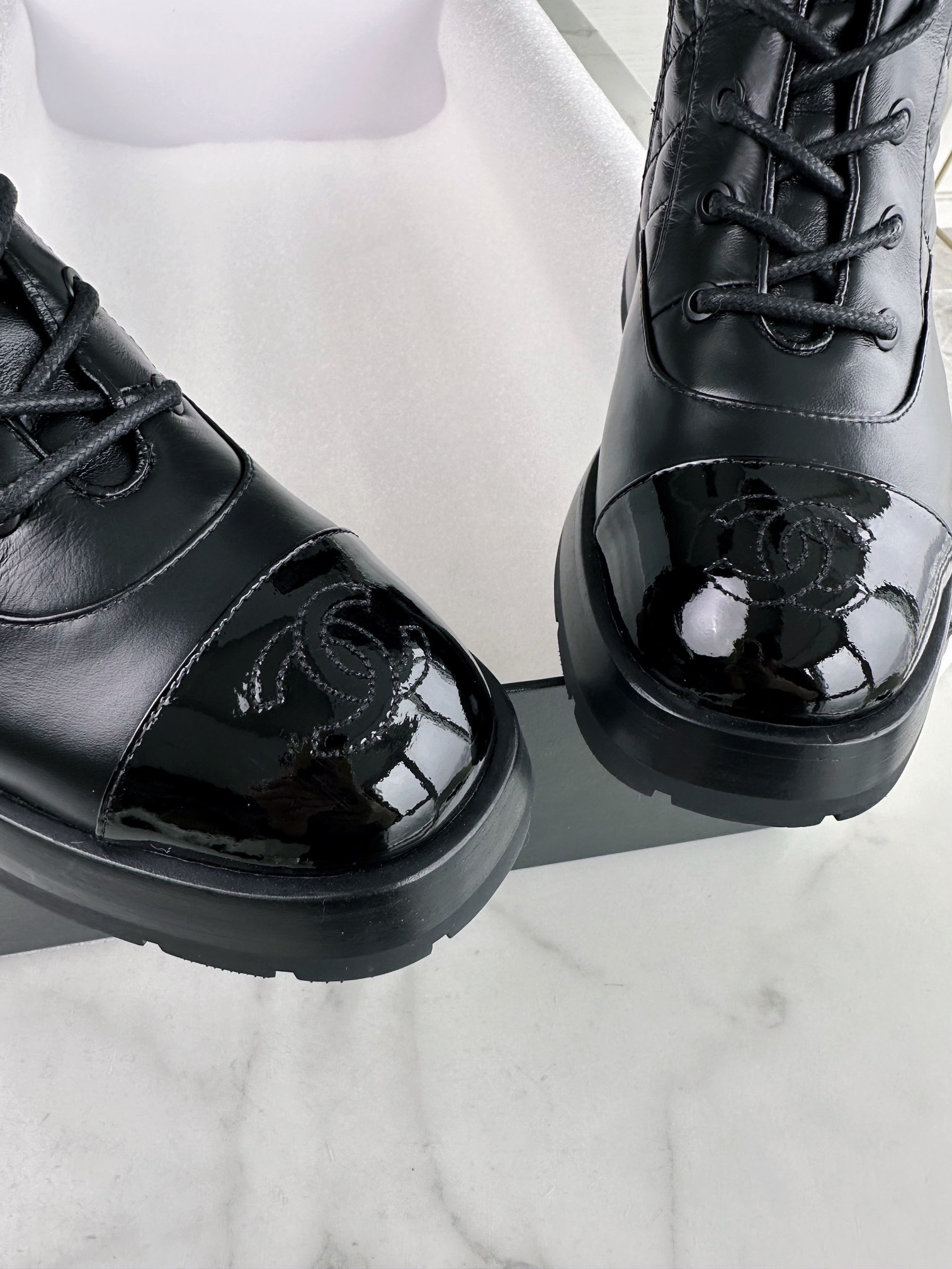 Chanel  23b  Chain Long Martin Boots: Crafted from black oil leather and black patent leather, with a leather combination sole. Classic diamond-pattern stitching and chain elements intertwine on the vamp